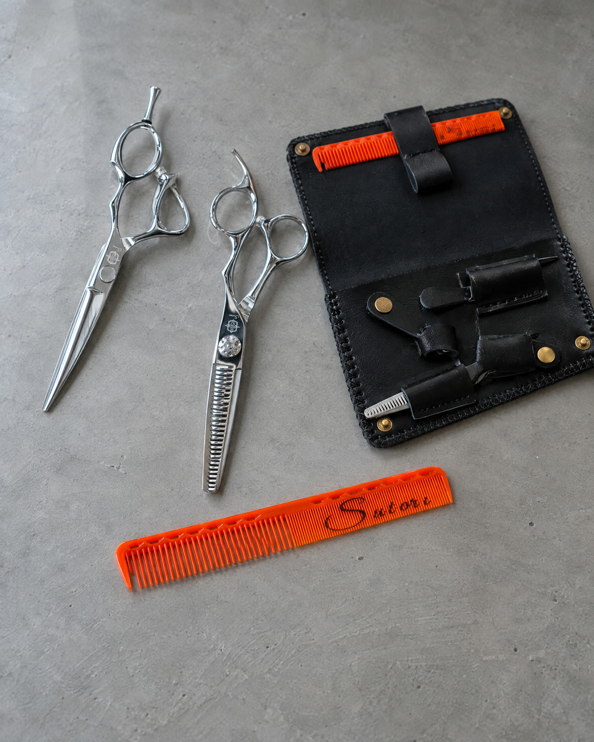 Sutori scissors, the double leather case and the orange cutting comb on grey microcement